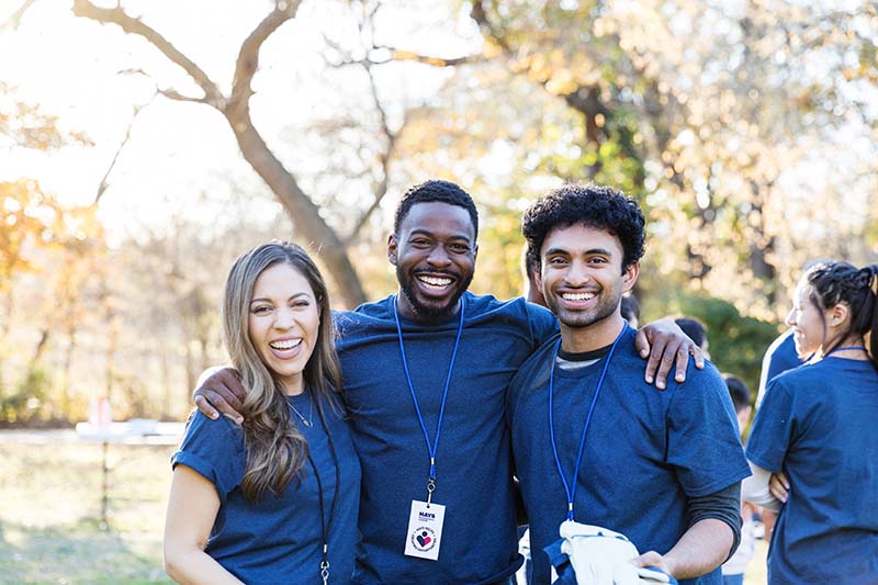 Drie personen dragen hetzelfde blauwe t-shirt en sleutelkoorden om hun nek. Ze glimlachen stralend voor een groepsfoto.  Het is een buitensetting met bomen op de achtergrond.
