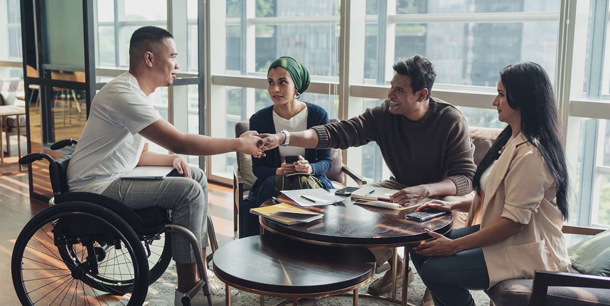 Een groep mensen zit rond een tafel en gaat met elkaar in gesprek. Er liggen blocnotes en pennen op tafel. Twee mannen schudden elkaar de hand over de tafel heen, één van hen zit in een rolstoel.