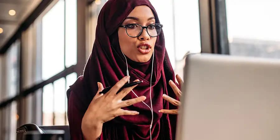 Een vrouw met een bril en een paarse hijab ziet er geanimeerd uit terwijl ze praat in een videogesprek op haar laptop.