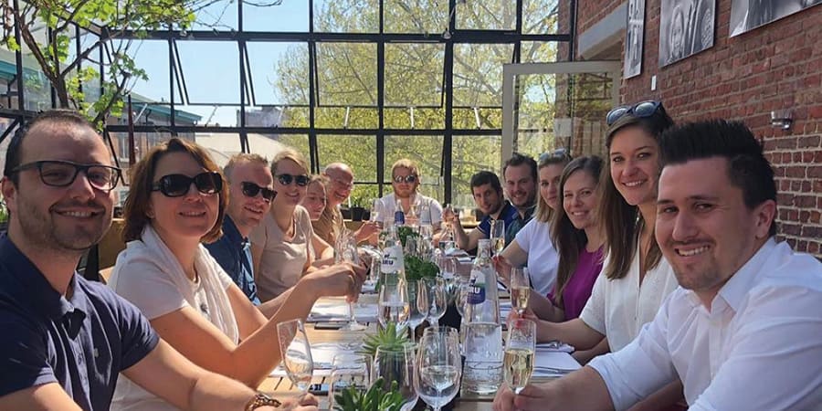 Een groep van ongeveer vijftien mensen zit aan een lange tafel iets te drinken. Ze glimlachen allemaal naar de camera. Ze dragen casual kleding.